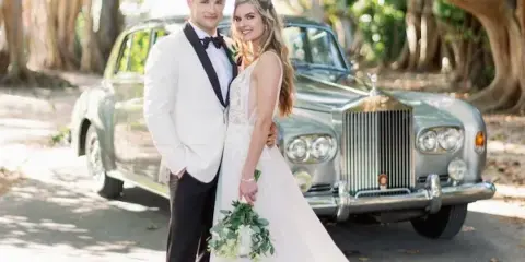 A bride and groom standing in front of a silver vintage Rolls-Royce car outdoors.