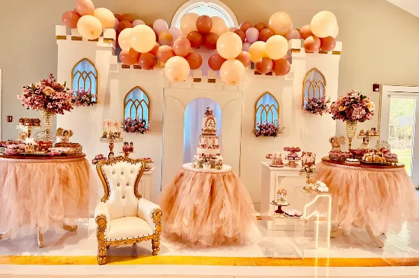 A luxury event decoration featuring a pastel balloon arch, floral arrangements, and a decorated dessert table with a tiered cake.
