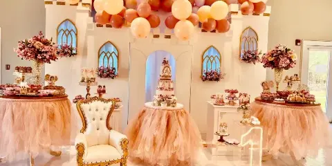 A luxury event decoration featuring a pastel balloon arch, floral arrangements, and a decorated dessert table with a tiered cake.
