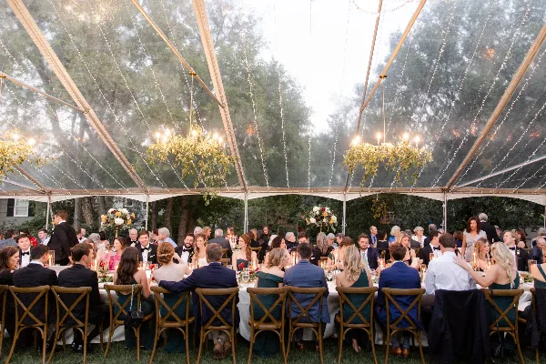 Outdoor wedding reception under a clear tent with string lights, floral arrangements, and seated guests.