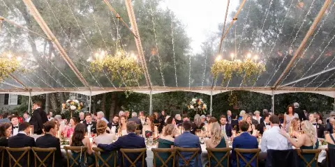 Outdoor wedding reception under a clear tent with string lights, floral arrangements, and seated guests.