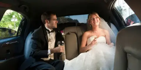 A bride and groom sitting in the back seat of a limousine holding champagne glasses.