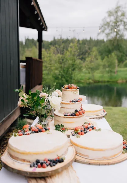 A multi-tiered wedding cake decorated with fresh berries and flowers, displayed on a table outdoors near a pond and a wooden lodge.