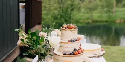 A multi-tiered wedding cake decorated with fresh berries and flowers, displayed on a table outdoors near a pond and a wooden lodge.