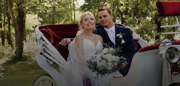 A bride and groom smiling while seated in a white vintage carriage with red velvet interior, holding a white floral bouquet.