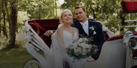 A bride and groom smiling while seated in a white vintage carriage with red velvet interior, holding a white floral bouquet.