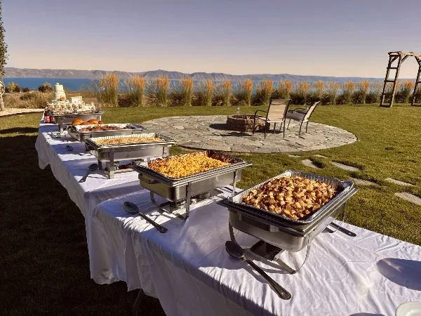 Outdoor catering buffet with multiple chafing dishes of food set up on a white table near a lake.