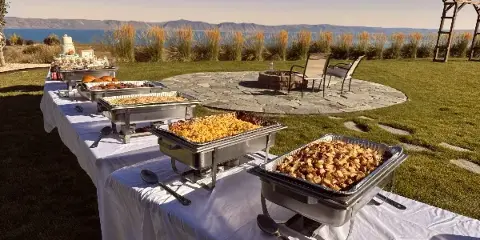 Outdoor catering buffet with multiple chafing dishes of food set up on a white table near a lake.