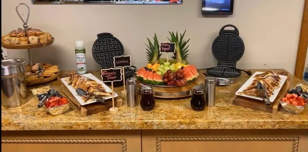 A catering buffet table featuring a large fruit platter, waffle makers, pastries, and prepared food trays.