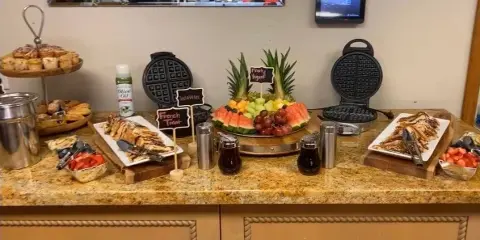 A catering buffet table featuring a large fruit platter, waffle makers, pastries, and prepared food trays.