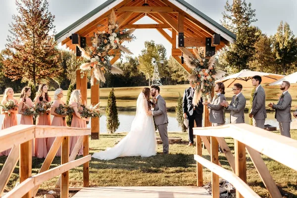 A wedding ceremony in progress at an outdoor wooden arbor venue with a lake and windmill in the background.