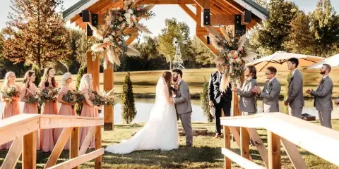 A wedding ceremony in progress at an outdoor wooden arbor venue with a lake and windmill in the background.