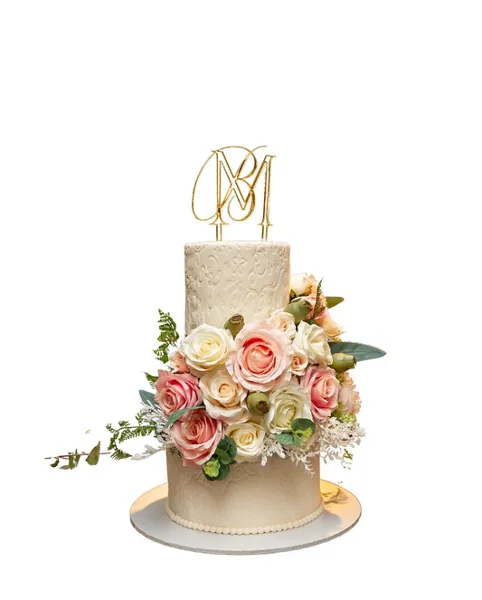 A two-tiered white wedding cake decorated with pink and white roses, greenery, and a gold monogram topper.