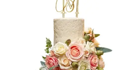 A two-tiered white wedding cake decorated with pink and white roses, greenery, and a gold monogram topper.