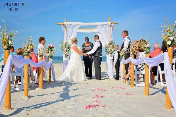 A bride and groom exchanging vows under a decorated arch on a sandy beach with seated guests.