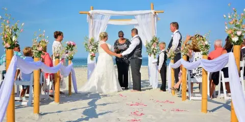 A bride and groom exchanging vows under a decorated arch on a sandy beach with seated guests.
