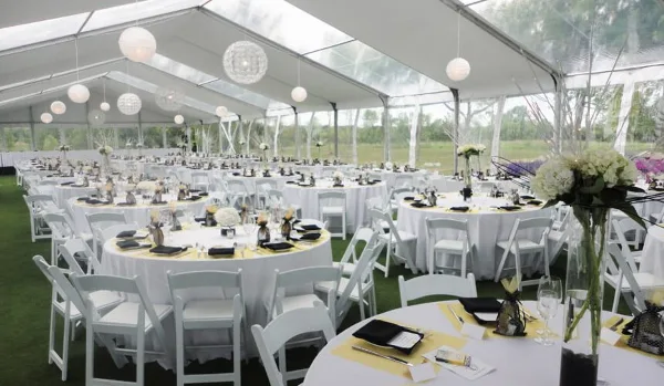 Interior of a clear span tent set up for a formal event with round tables, white chairs, and hanging globe lights.