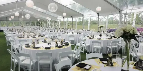 Interior of a clear span tent set up for a formal event with round tables, white chairs, and hanging globe lights.
