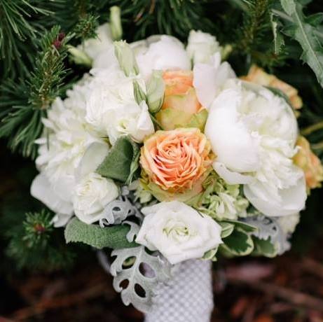 A close-up photograph of a floral bouquet featuring white and peach roses, white hydrangeas, and greenery.
