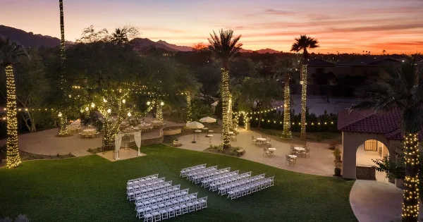 An outdoor wedding venue at dusk featuring rows of white chairs, a decorated arch, and palm trees wrapped in string lights.