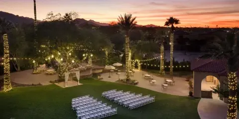 An outdoor wedding venue at dusk featuring rows of white chairs, a decorated arch, and palm trees wrapped in string lights.