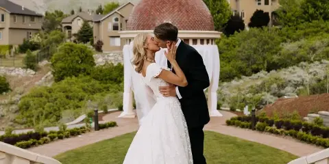 A bride and groom sharing a kiss in front of a white wedding arch in a landscaped garden setting.