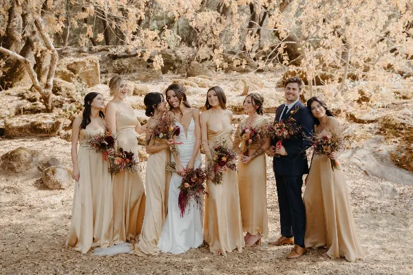 A wedding party consisting of a bride, groom, and attendants posing together in a wooded area with dry leaves and oak trees.