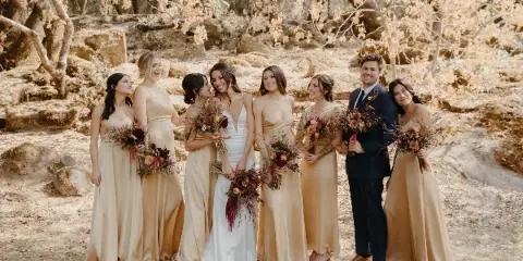 A wedding party consisting of a bride, groom, and attendants posing together in a wooded area with dry leaves and oak trees.