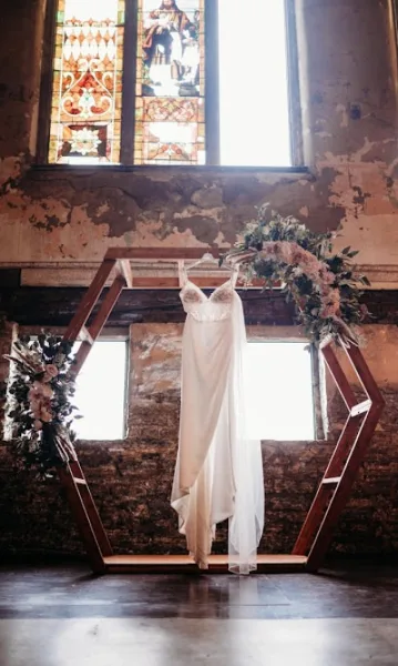 A white wedding dress hanging on a wooden frame decorated with floral arrangements in a rustic venue.