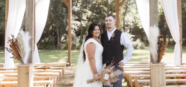 A bride and groom standing in a rustic open-air wooden wedding chapel with white drapery and pampas grass arrangements.