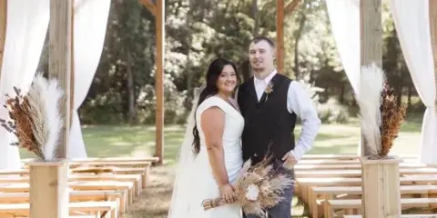 A bride and groom standing in a rustic open-air wooden wedding chapel with white drapery and pampas grass arrangements.