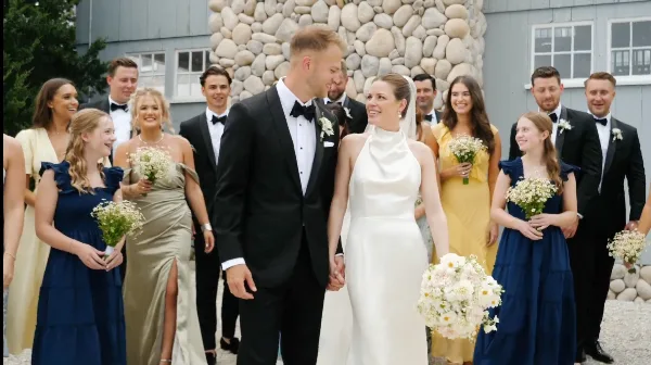 A wedding party consisting of the bride, groom, and attendants posing together outdoors in front of a stone wall.