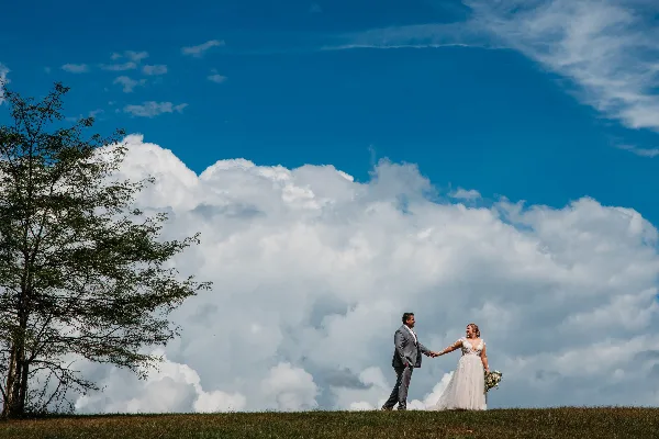 A bride and groom holding hands on a grassy hill against a blue sky with large white clouds.