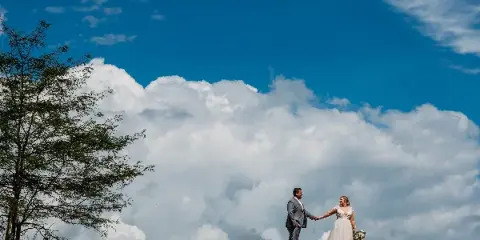 A bride and groom holding hands on a grassy hill against a blue sky with large white clouds.