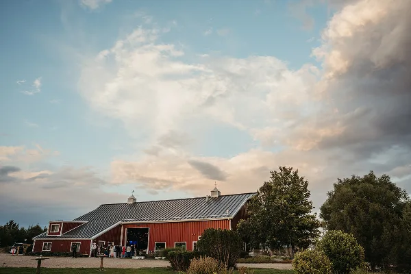 Exterior view of a large red barn event venue with a metal roof, set against a cloudy sky with trees and landscaping in the foreground.