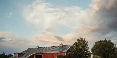 Exterior view of a large red barn event venue with a metal roof, set against a cloudy sky with trees and landscaping in the foreground.