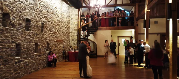 Wedding couple dancing at a reception in a rustic stone-walled venue with guests and a balcony overlooking the dance floor.