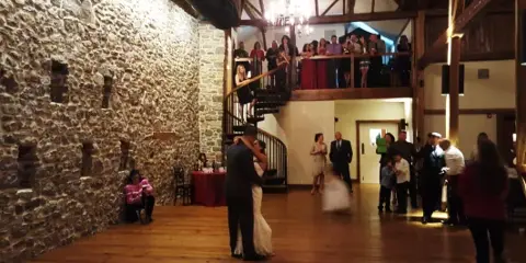 Wedding couple dancing at a reception in a rustic stone-walled venue with guests and a balcony overlooking the dance floor.