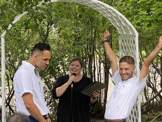 A wedding officiant speaks into a microphone while officiating a ceremony for two couples standing under a white arch.