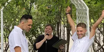 A wedding officiant speaks into a microphone while officiating a ceremony for two couples standing under a white arch.