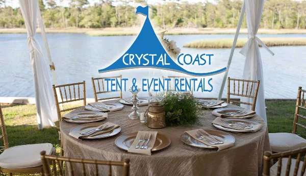 A set round event table with gold chiavari chairs, linens, and flatware under a white tent near a body of water.