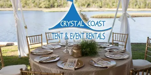 A set round event table with gold chiavari chairs, linens, and flatware under a white tent near a body of water.