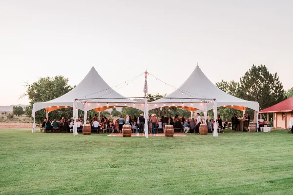A large white event tent with multiple peaks set up on a grassy field, illuminated by string lights, with guests seated inside.