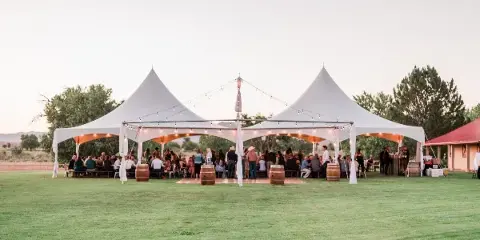 A large white event tent with multiple peaks set up on a grassy field, illuminated by string lights, with guests seated inside.