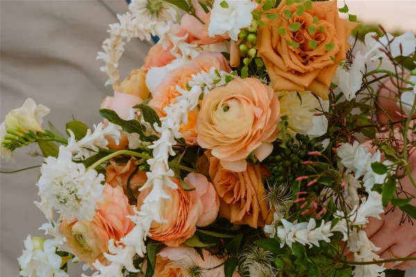 Close-up of a hand-held wedding bouquet featuring peach roses, ranunculus, and white flowers