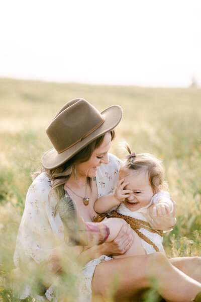 A mother wearing a hat holds a smiling toddler in a grassy field.