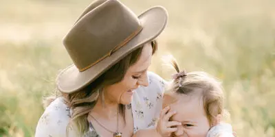 A mother wearing a hat holds a smiling toddler in a grassy field.