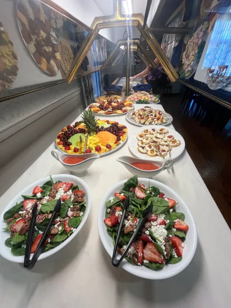 Buffet table with spinach strawberry salad, fruit platter, and assorted appetizers with serving tongs