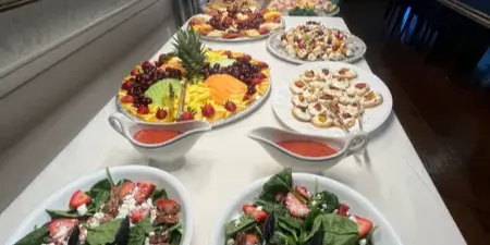 Buffet table with spinach strawberry salad, fruit platter, and assorted appetizers with serving tongs
