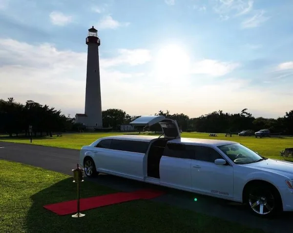 A white stretch limousine parked on a paved path with a red carpet leading to the open door, set against a backdrop of a lighthouse and green grass.